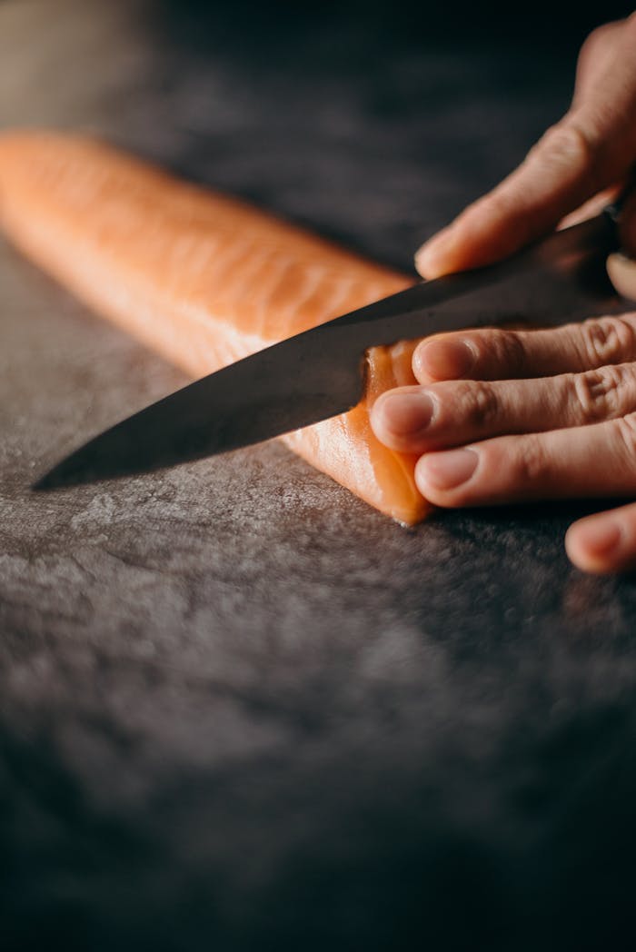 A sharp knife slicing fresh salmon fillet on a dark surface, showcasing food preparation skills.